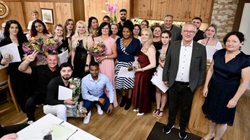 Eine Gruppe von Menschen, von denen einige Blumen und Geschenke in der Hand halten, posiert und lächelt in einem warm beleuchteten Raum mit holzgetäfelten Wänden, was auf ein feierliches Ereignis oder eine Zusammenkunft hindeutet.