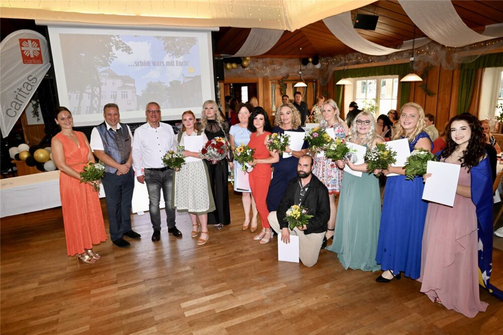Eine Gruppe formell gekleideter Männer und Frauen steht im Gebäude. Einige halten Blumensträuße und Urkunden in den Händen und lächeln in die Kamera. Hinter ihnen sind Luftballons, ein Banner der Caritas Berufsfachschule für Pflege Landshut und eine Projektionsfläche zu sehen.