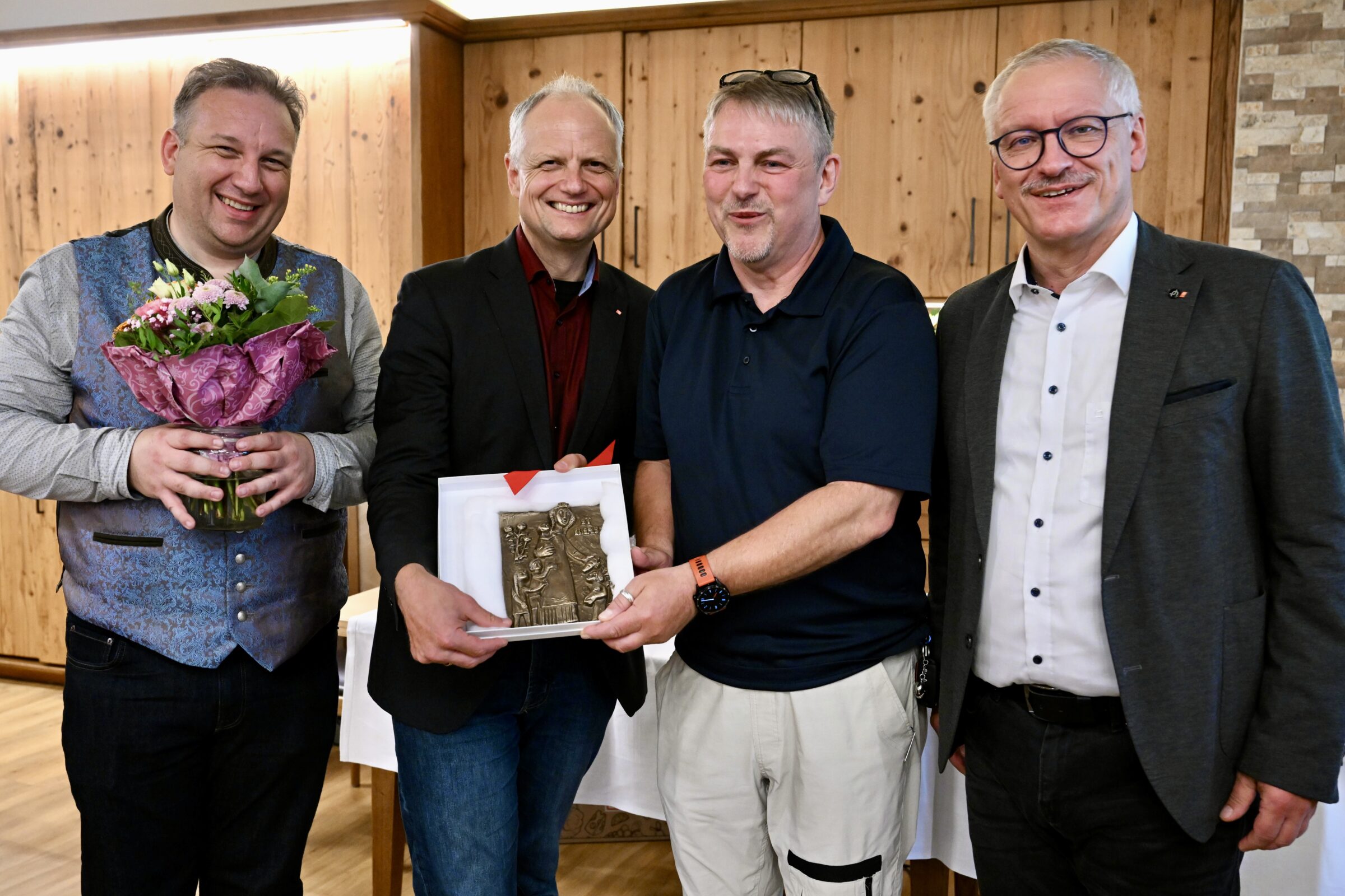 Vier lächelnde Männer stehen in einem Haus; einer hält einen Blumenstrauß, ein anderer überreicht eine Plakette oder eine Auszeichnung. Sie sind halb-formal gekleidet, und es scheint sich um eine Feier oder Anerkennung zu handeln.