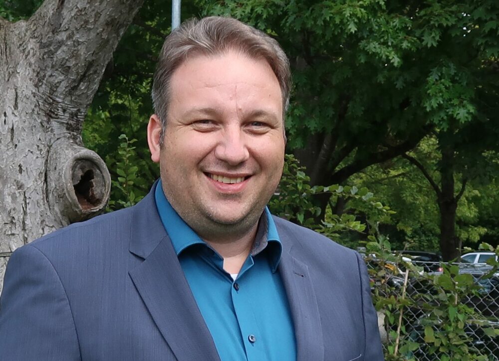 Ein Mann in einem blauen Jackett und einem blaugrünen Hemd lächelt im Freien vor einem Baum und grünem Laub, im Hintergrund sind ein Maschendrahtzaun und geparkte Autos zu sehen.
