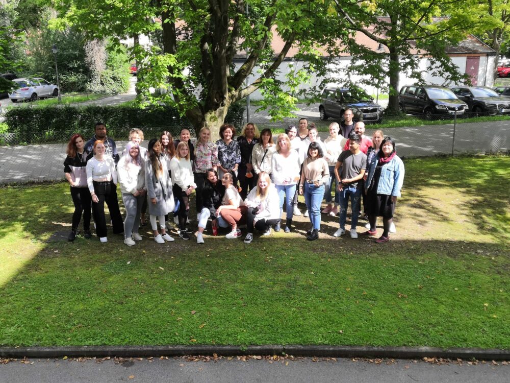 Eine Gruppe von etwa 30 Personen steht gemeinsam auf einer Rasenfläche unter einem großen Baum und lächelt in die Kamera. Hinter ihnen parken Autos, ein Zaun und einige Gebäude, durch deren Blätter das Sonnenlicht fällt.