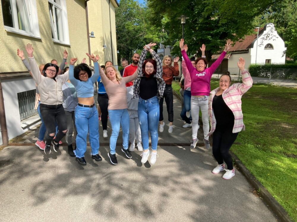 Eine Gruppe von Menschen im Freien, die an einem sonnigen Tag lächeln und begeistert in die Luft springen, vor einem gelben Gebäude mit Bäumen und Gras im Hintergrund.