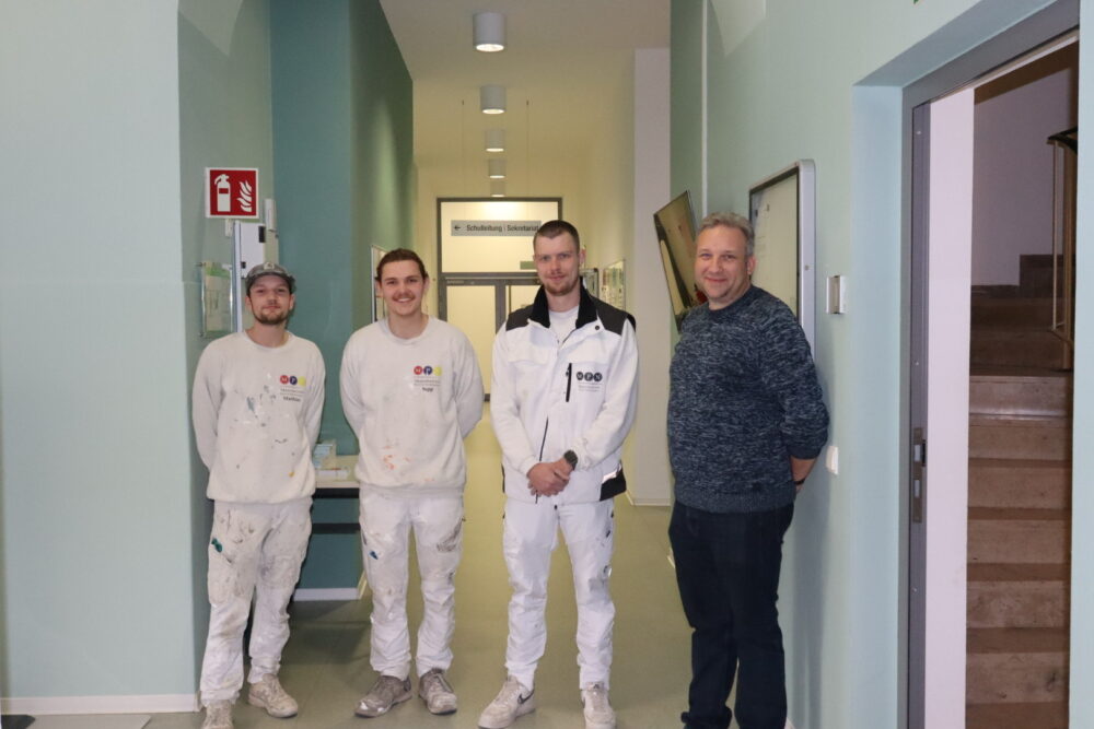 Vier Männer stehen in einem Flur. Drei tragen weiße, mit Farbe bespritzte Maleruniformen, einer trägt einen blauen Pullover und eine schwarze Hose. Sie lächeln und blicken in die Kamera; im Hintergrund sind Schilder und Treppen zu sehen.