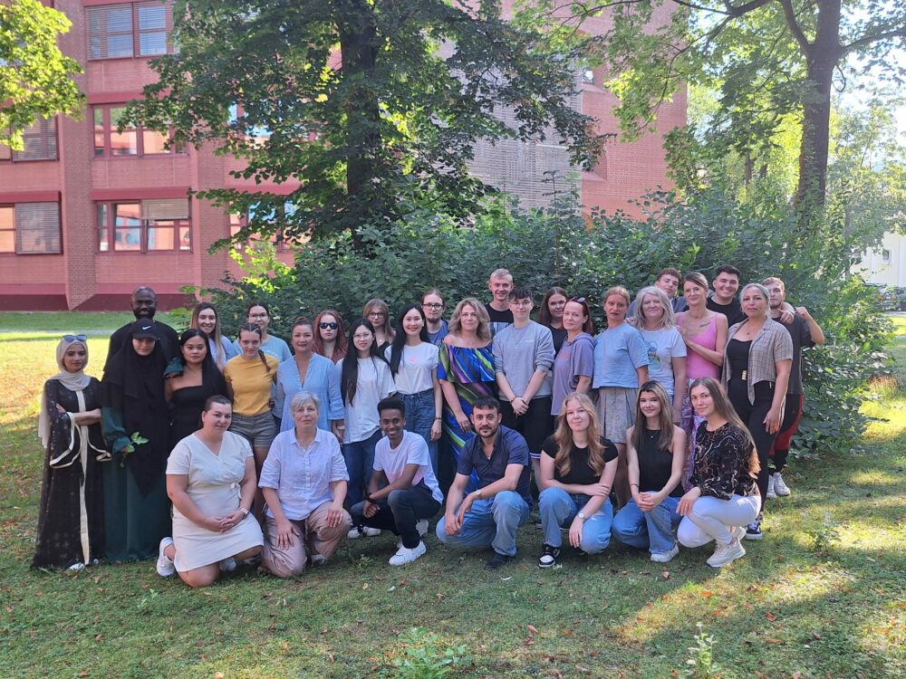 Eine große Gruppe von Menschen, die einen Pflegedienst in Landshut repräsentieren, posiert im Freien vor grünen Büschen und Bäumen, hinter ihnen ein rotes Backsteingebäude. Verschiedene Männer und Frauen stehen und knien im Gras und lächeln in die Kamera.