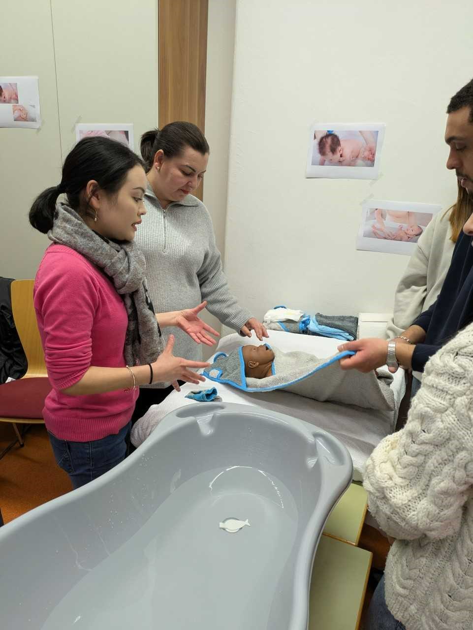 Vier Erwachsene stehen um eine in ein Handtuch gewickelte Babypuppe herum und üben neben einer grauen Babybadewanne Badetechniken. An der Wand hinter ihnen hängen Poster mit Anweisungen zur Babypflege.