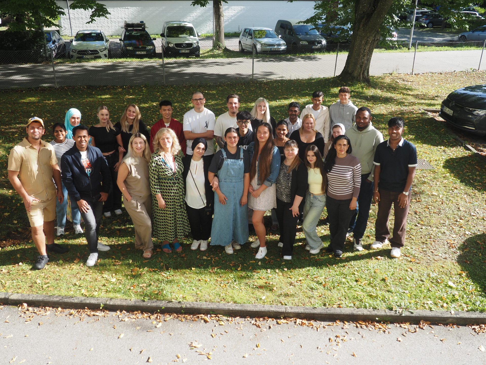 Eine Gruppe von etwa 28 Personen posiert an einem sonnigen Tag im Freien, auf einer Wiese mit Bäumen und geparkten Autos im Hintergrund. Die meisten lächeln und schauen in die Kamera.